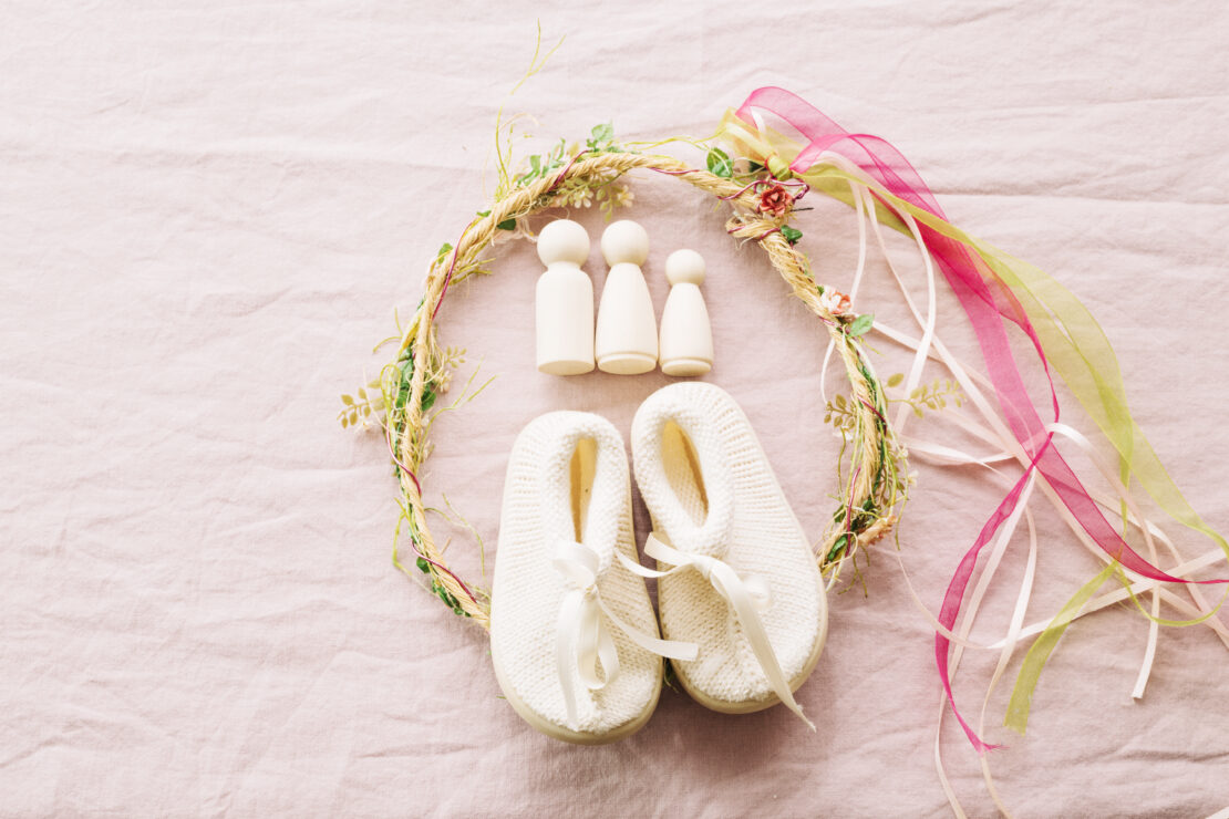 Petits chaussons de bébé blancs posés sur un tissu rose, entourés d’une couronne florale et de rubans colorés